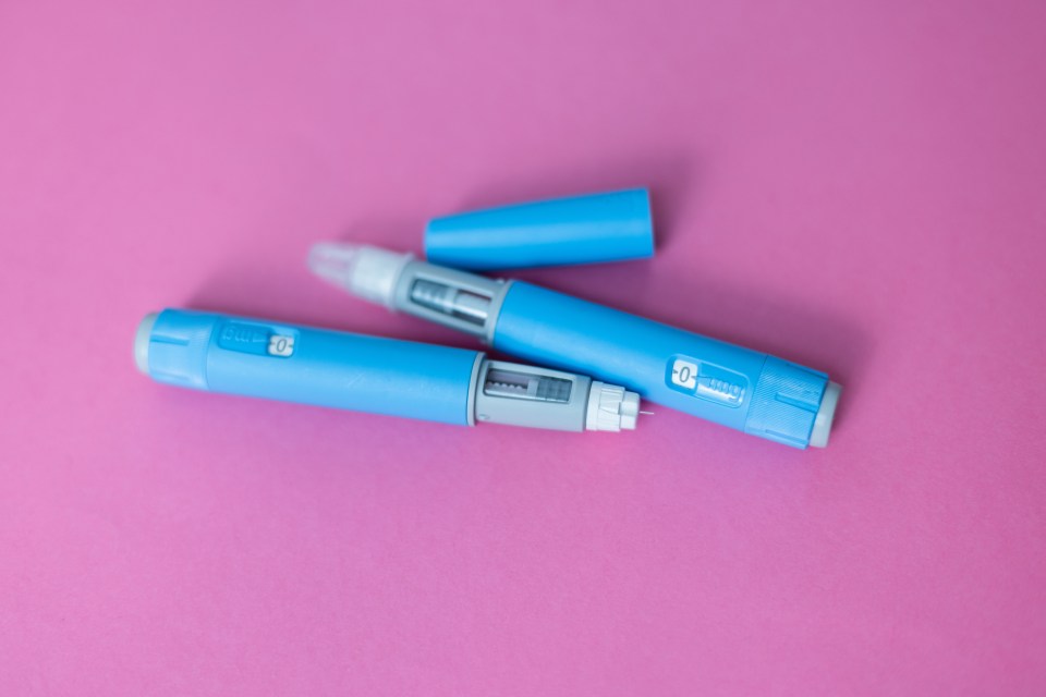Two blue insulin pens, one capped and one uncapped with a needle exposed, on a pink background.