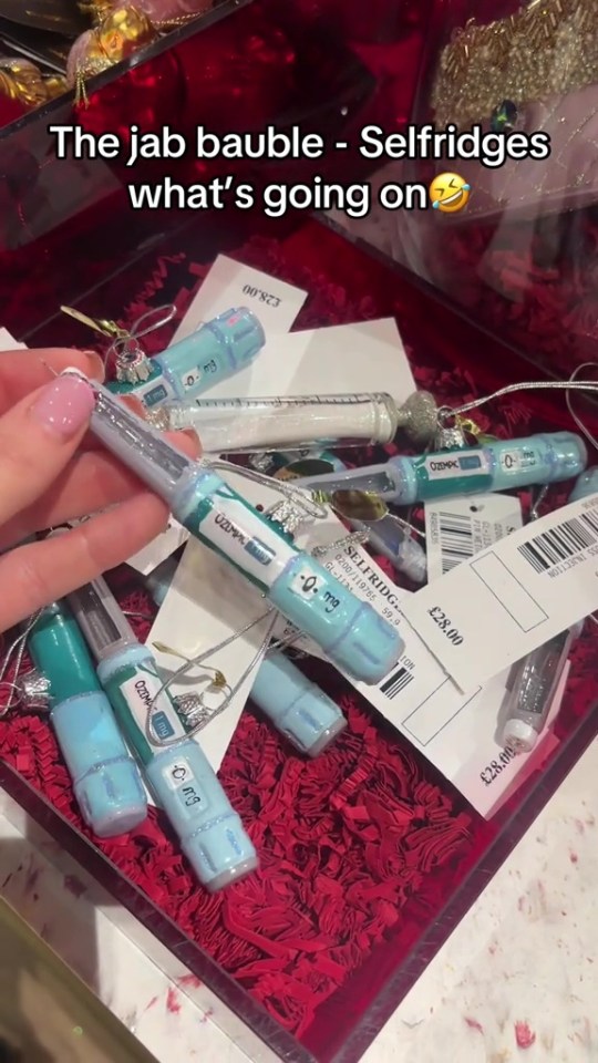 Hand holding an Ozempic pen-shaped Christmas ornament over a box filled with similar ornaments and price tags.