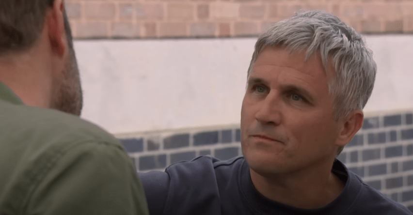 A man with gray hair in a blue shirt looks toward another man from behind.
