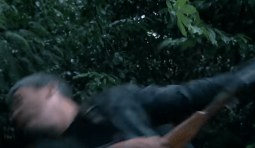 Man in a black jacket and gray hair being thrown or pushed back with dark foliage in the background.