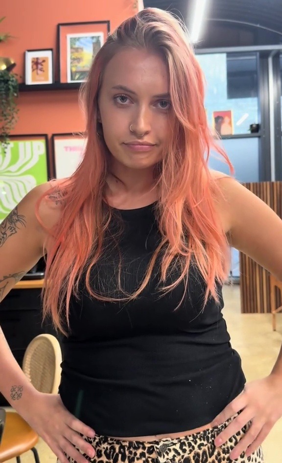 A woman with light pink hair and tattoos on her arms, wearing a black tank top and leopard print pants, stands with her hands on her hips.