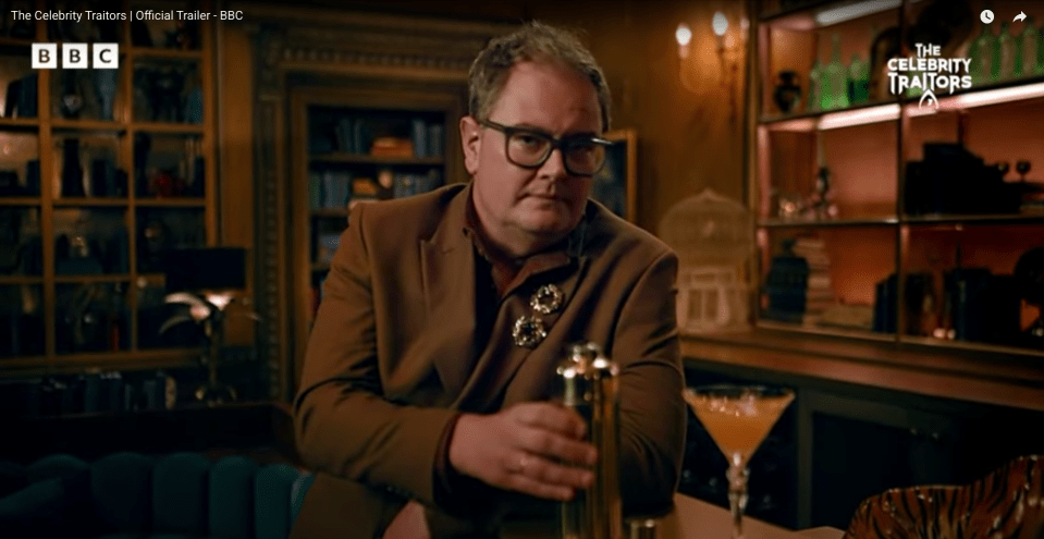 Alan Carr in a brown suit and glasses holding a cocktail shaker.