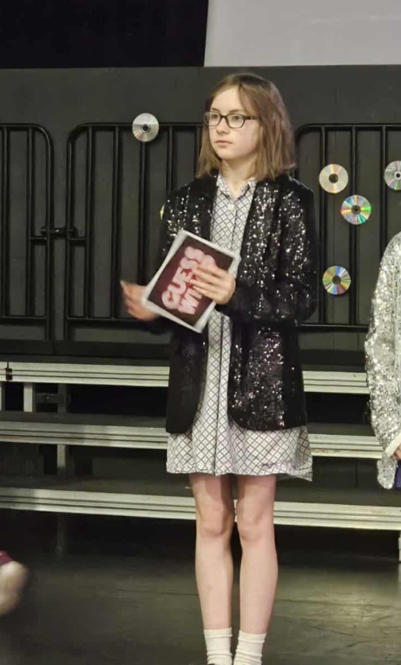 Sienna in a black sequined jacket, patterned dress, and black Mary Jane shoes holding a book that says "GUESS WHO" on the cover.