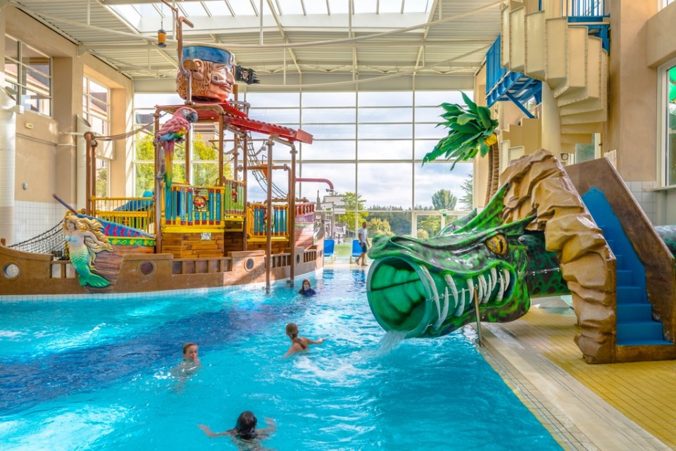 Indoor swimming pool with children playing near a pirate ship and a green dragon slide.