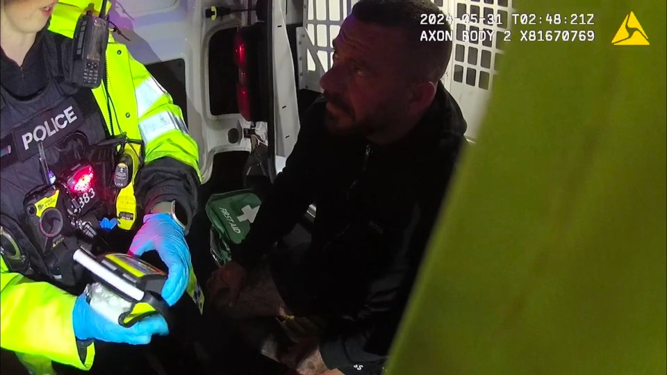 A police officer in a high-visibility jacket with the word "POLICE" on her vest, speaking to a man inside the back of a police van. The officer is holding a device while wearing blue gloves.