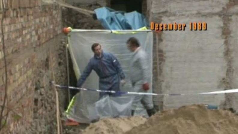 Paul Bodenham in a blue jumpsuit stands next to another man in white inside a makeshift tent, with a pile of sand in the foreground.