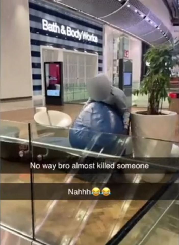 A person in a gray hoodie pushing a large blue beanbag chair over a glass barrier in a shopping center, with a Bath & Body Works store in the background.