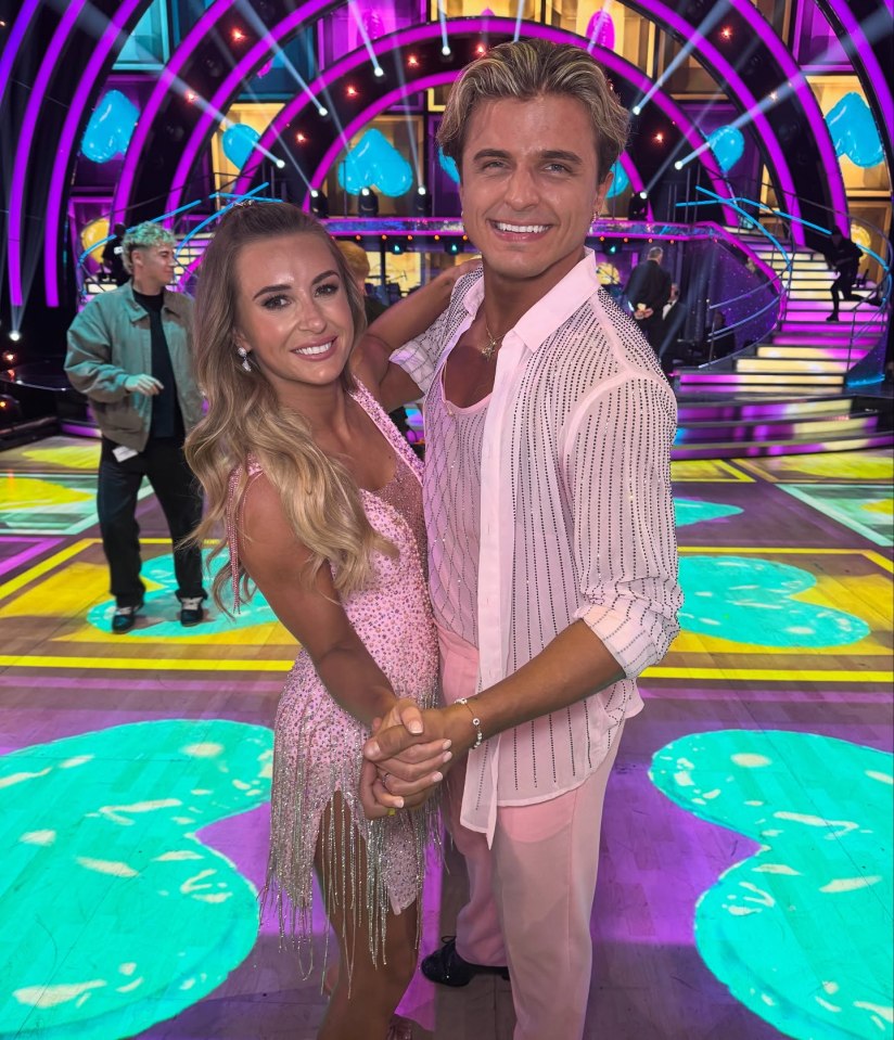 Nikita Kuzmin and Dani Dyer smiling and holding hands in pink outfits on a dance floor with a lit-up stage behind them.