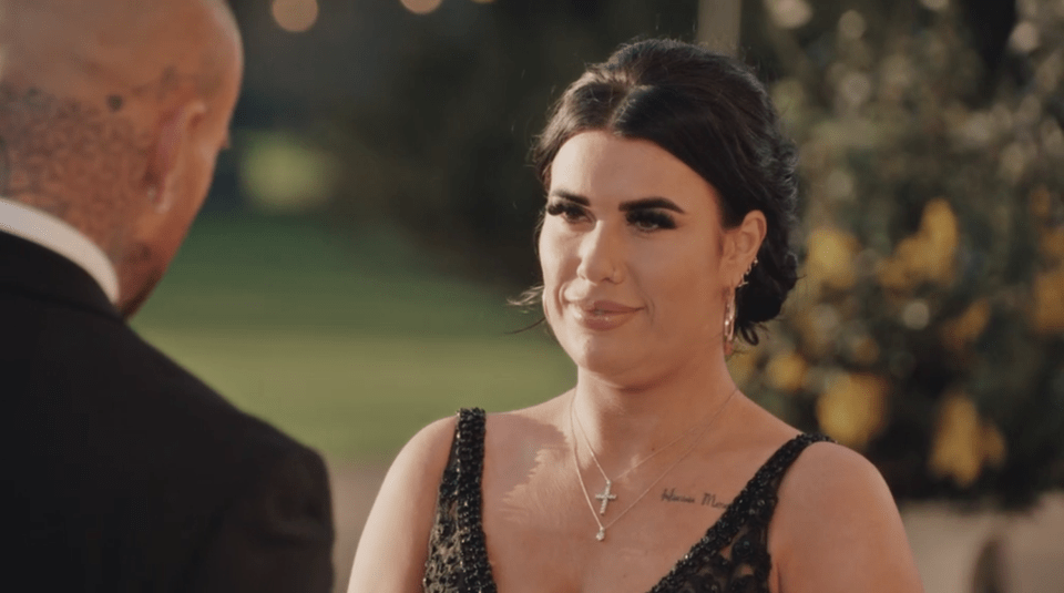 A woman in a black dress with a cross necklace smiling at a man in a suit.