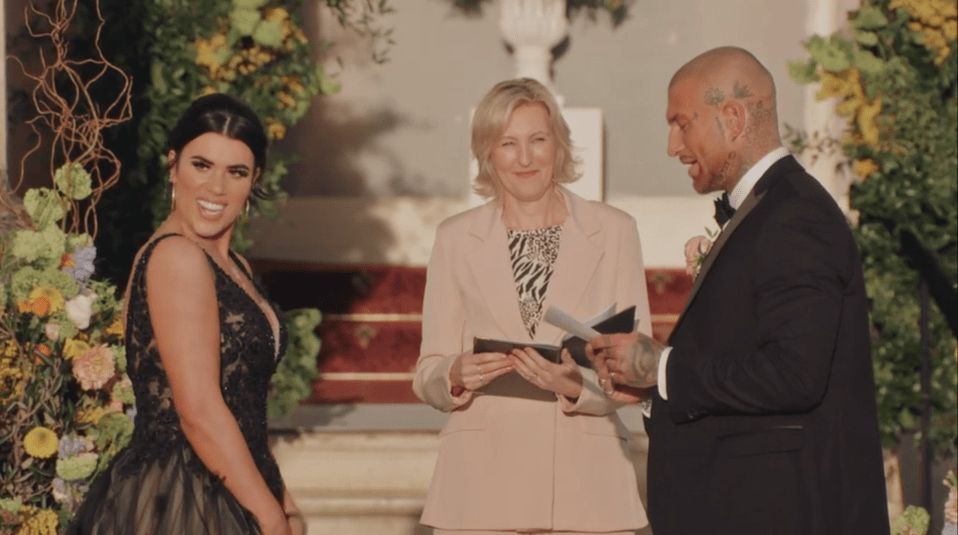 A woman in a black gown looking at the officiant who is holding a book, while the man in a suit looks at her.