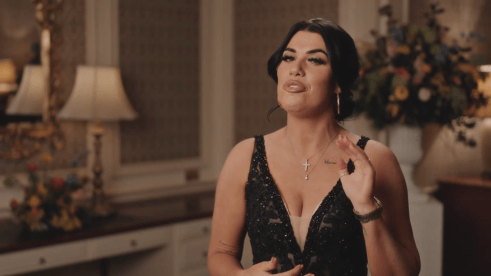 A woman in a black dress with a cross necklace, talking and gesturing.