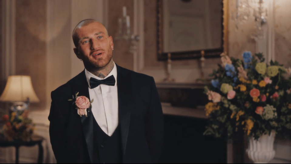 A man in a tuxedo with a pink boutonniere and tattoos on his neck and arm smiles and looks up.