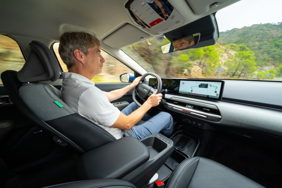 A man driving a Kia car on a winding road with mountains in the background.