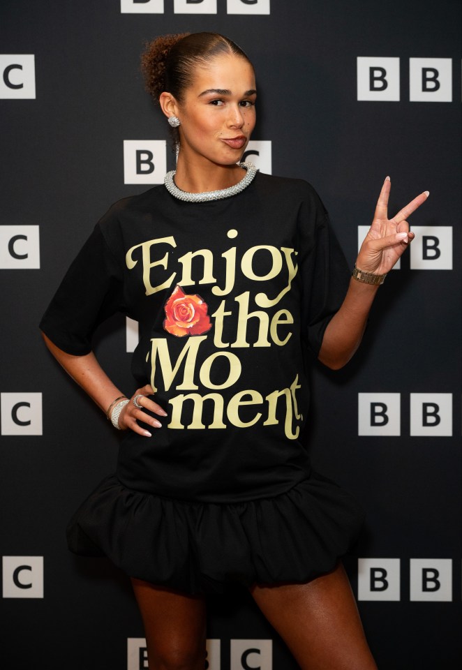 Tillie Amartey posing in a black graphic print mini dress with the text "Enjoy the Moment" and a red rose, while making a peace sign at the "Glow Up" Series Seven Screening.