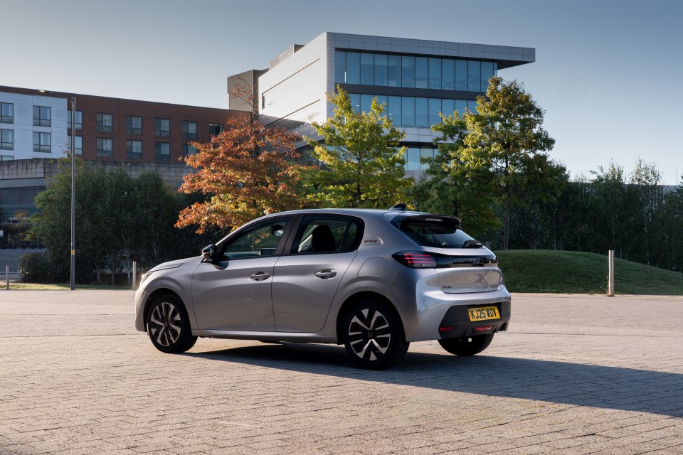 A silver Peugeot 208 parked on a paved lot in front of modern buildings and trees with autumn foliage.