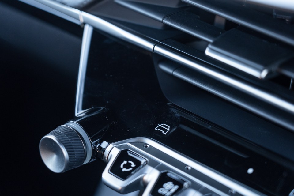 Close-up of a car's interior, showing part of the dashboard with air vents, a rotary knob, and buttons with icons, including one resembling a car.
