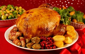 A roasted Christmas turkey dinner with brussels sprouts, broccoli, carrots, pigs in blankets, roasted potatoes, stuffing, and cranberries, on a red background.