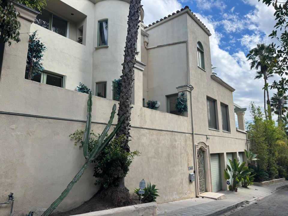 The exterior of a beige stucco home with a palm tree, cacti, and a security sign in front.