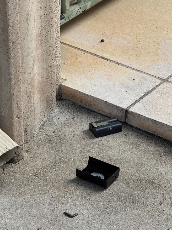 A black Ring doorbell battery and its empty casing lie on concrete ground next to a concrete wall.