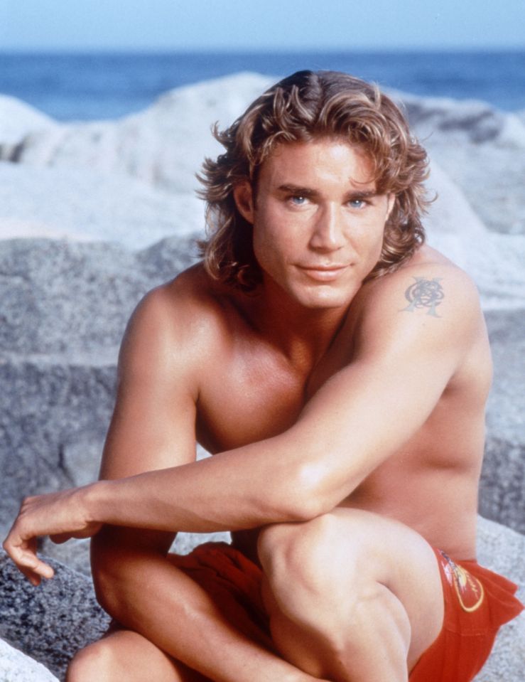 Jaason Simmons from 'Baywatch' in red trunks, smiling at the camera, with a tattoo on his left shoulder, sitting on rocks with the ocean in the background.