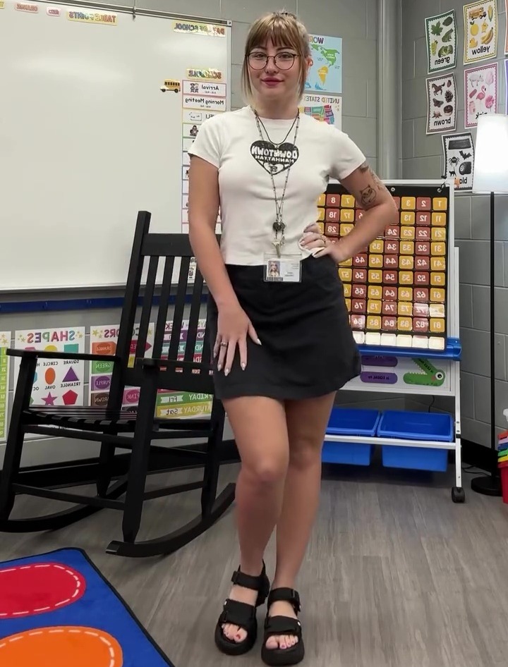 A teacher in a white t-shirt and black skirt standing in a classroom.