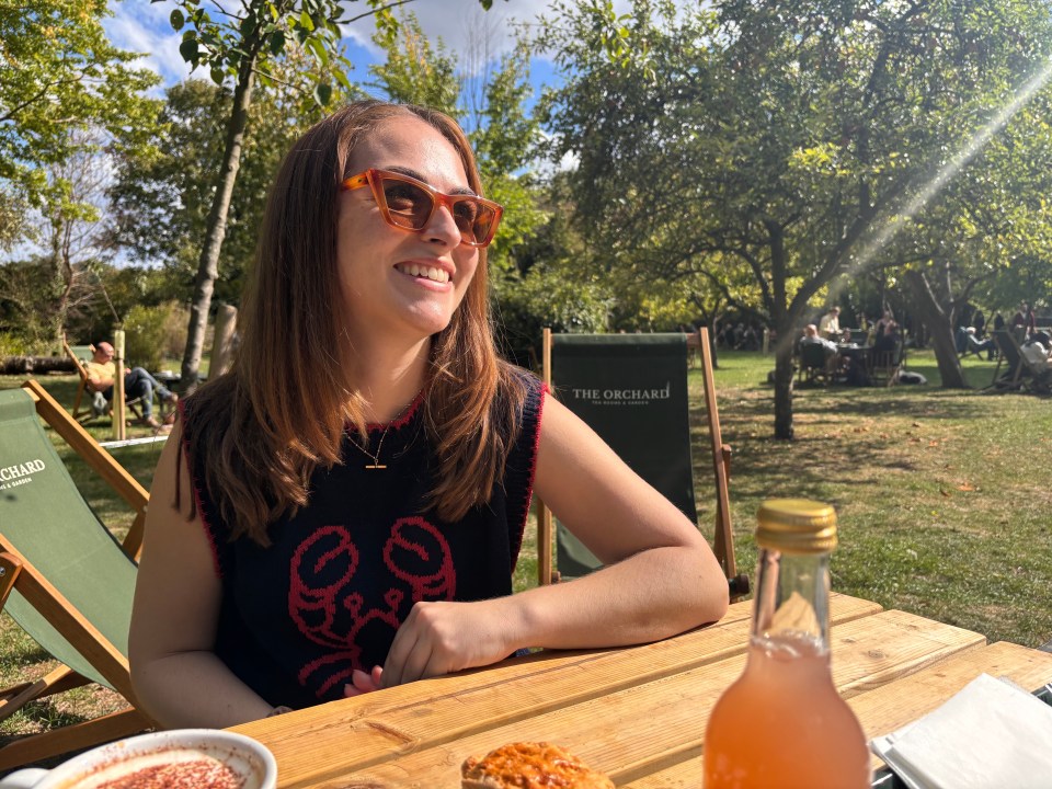 A young woman with brown hair and orange sunglasses sitting at a wooden table in an outdoor tea garden.