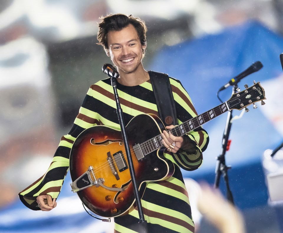 Harry Styles performing on stage, smiling and holding a guitar, wearing a black, neon green, and brown striped outfit.