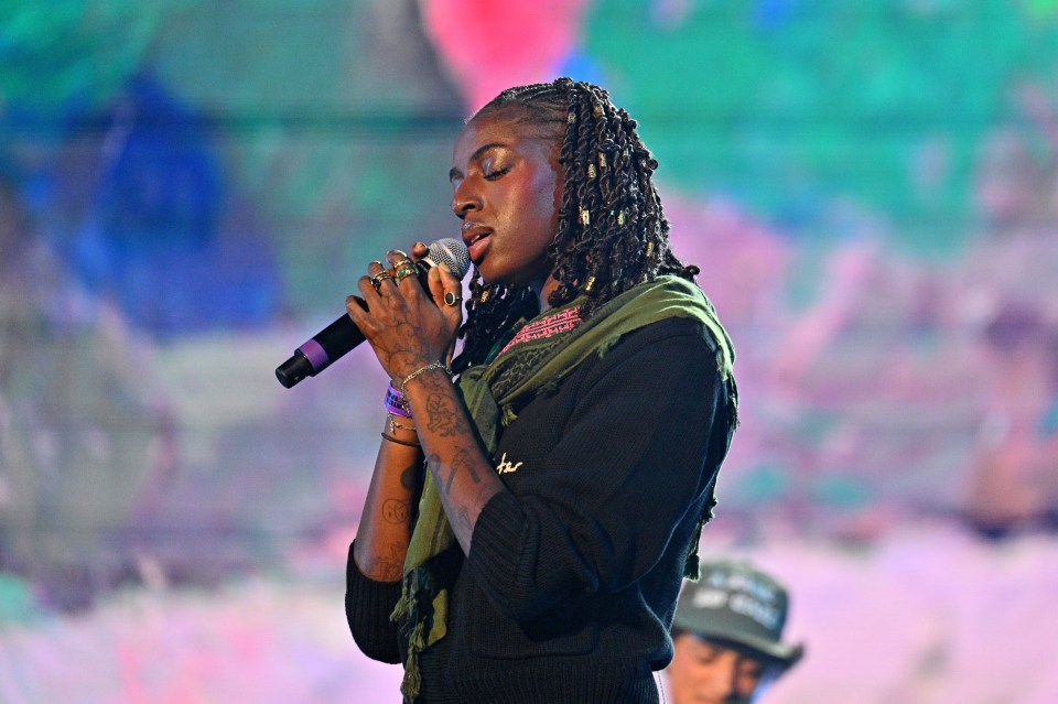 Cat Burns performing onstage, wearing a green scarf and black top, holding a microphone.