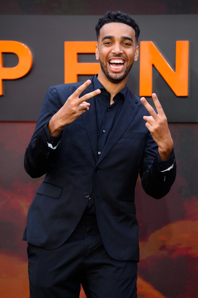 Niko Omilana attends the Oppenheimer UK Premiere, wearing a black suit and open-collar shirt, smiling and giving two peace signs.