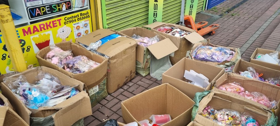 Cardboard boxes overflowing with counterfeit Labubu dolls and toys on a sidewalk outside a "VAPE SHOP."
