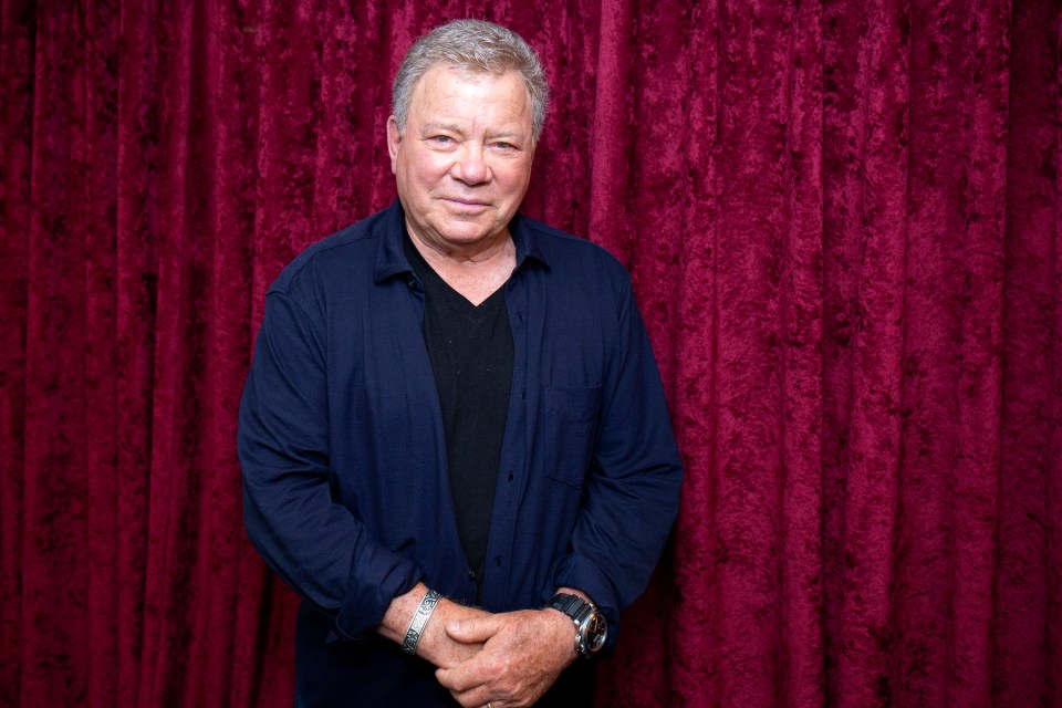 William Shatner in a dark blue shirt over a black t-shirt, standing in front of red velvet curtains.