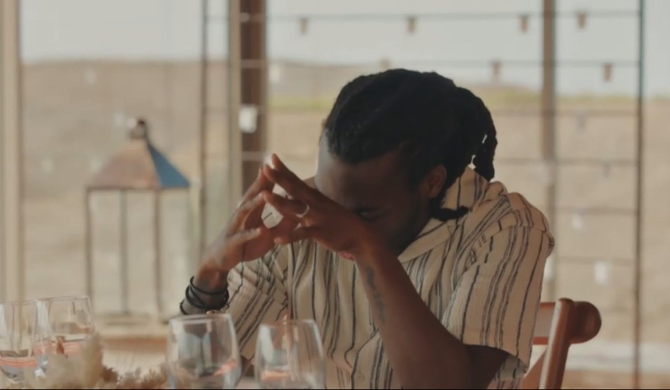 A man wearing a striped shirt sits at a table with his hands clasped over his face.
