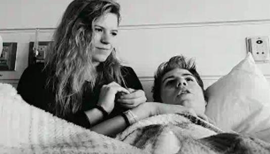 Sabia Reiche holding Loren Schauers' hand while he is lying in a hospital bed.