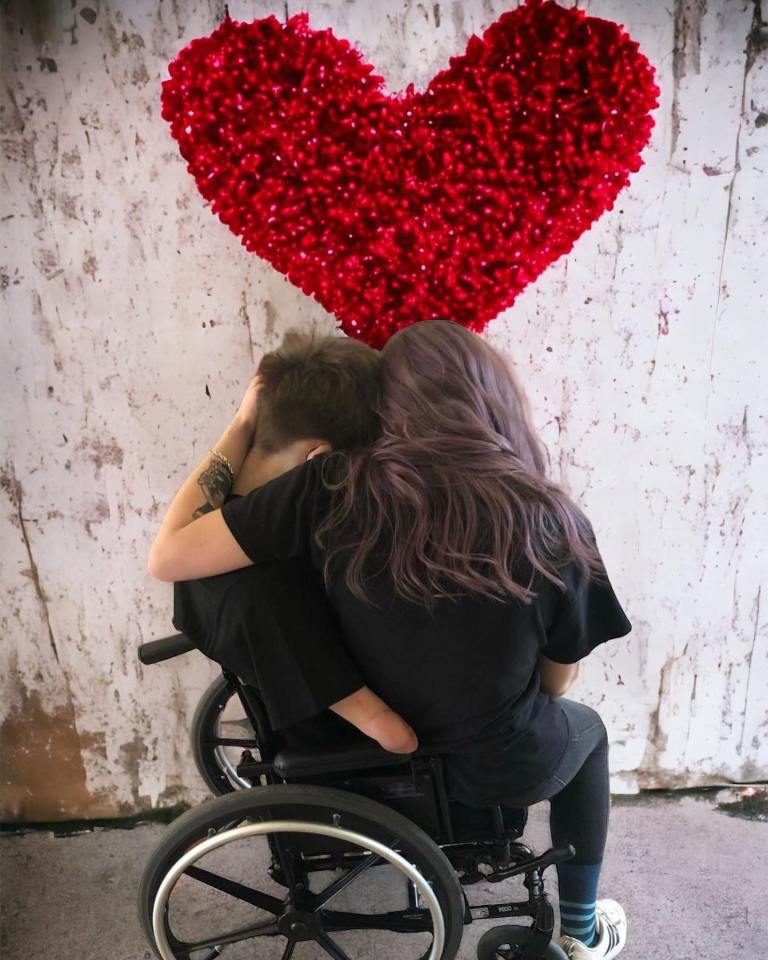 A woman sitting in a wheelchair embraces a man from behind, beneath a large red heart decoration.