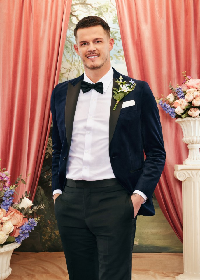 Steven from Married At First Sight, wearing a dark blue suit jacket with a black bow tie and white shirt, stands smiling in front of a draped coral background with floral arrangements.