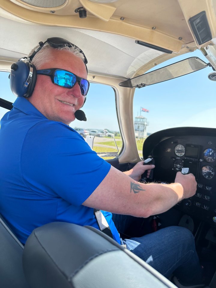 A man in a blue shirt, sunglasses, and headset smiling in a cockpit.