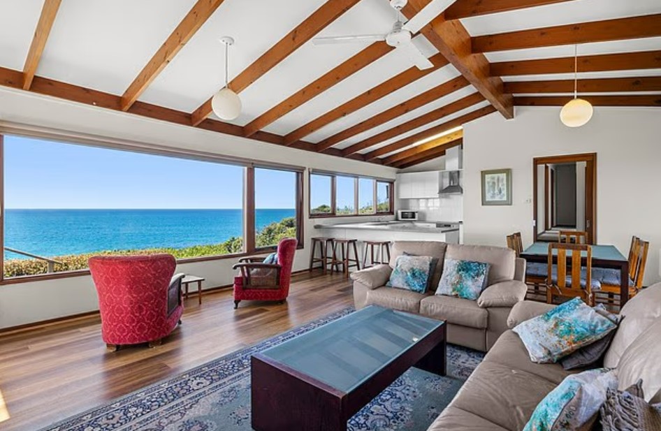 Interior of a house with an ocean view, showing a living room with two couches, a kitchen, and a dining area.