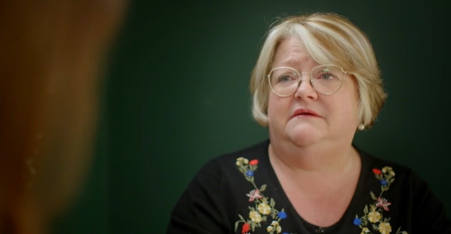 A woman wearing glasses and a black top with embroidered flowers looks emotional.