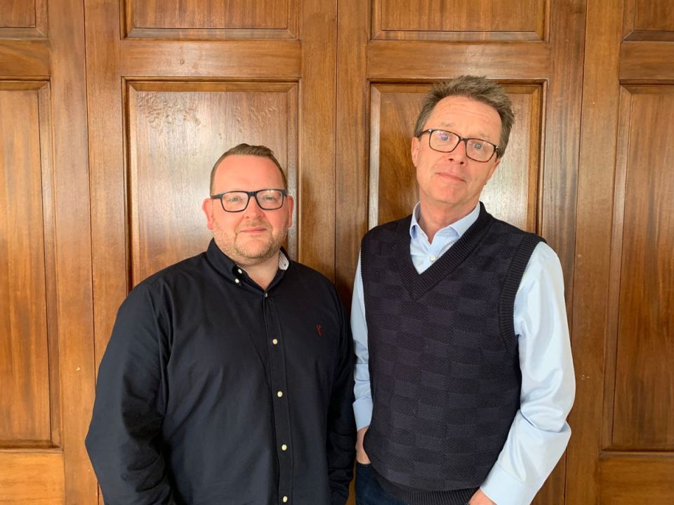 A man with glasses and a dark shirt standing next to another man with glasses, a light blue shirt, and a dark vest, in front of a wooden door.
