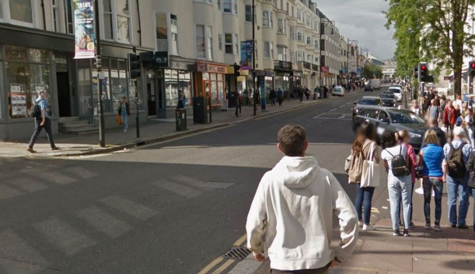 A busy street in Brighton with shops, cars, and pedestrians, viewed from behind a man in a white hoodie.