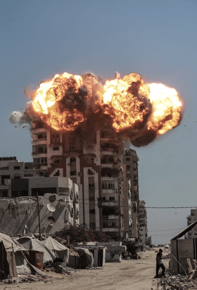 An explosion of fire and smoke erupts from the top of a multi-story building in a damaged urban landscape.
