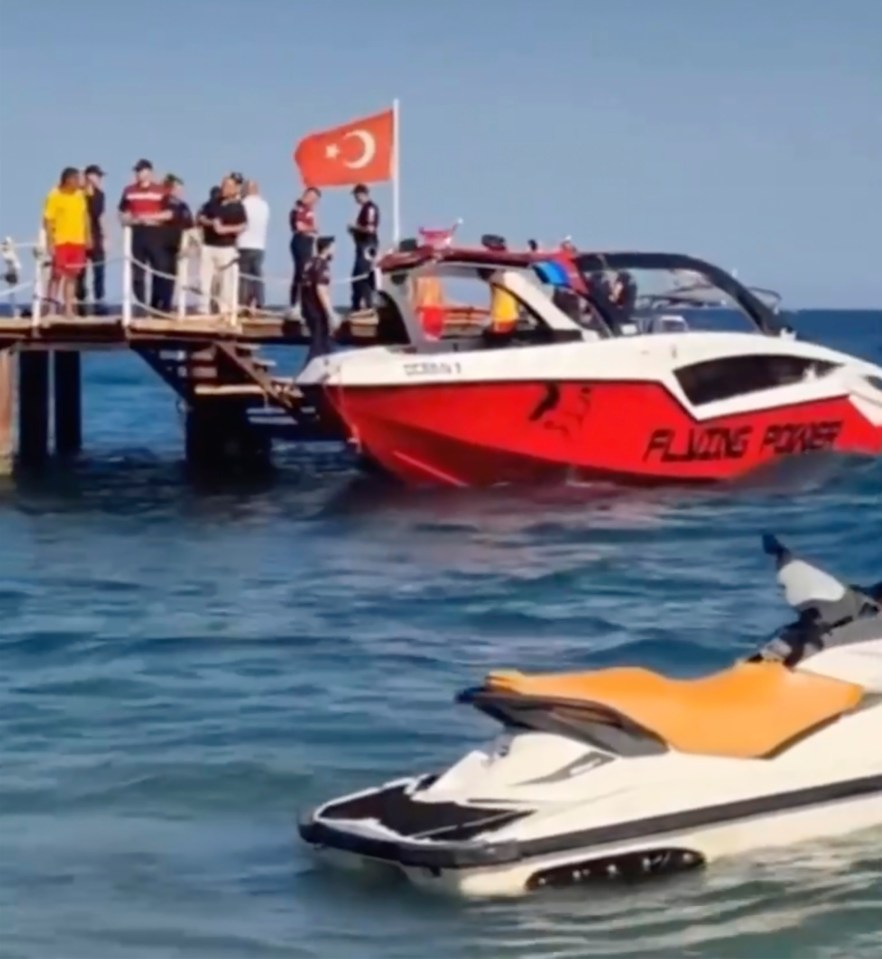 A group of people standing on a pier next to a red and white speed boat with a Turkish flag, and a jet ski in the water.