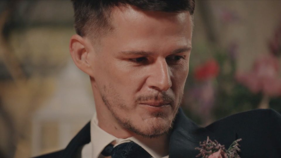 A groom with a beard and dark suit looks down with a solemn expression.