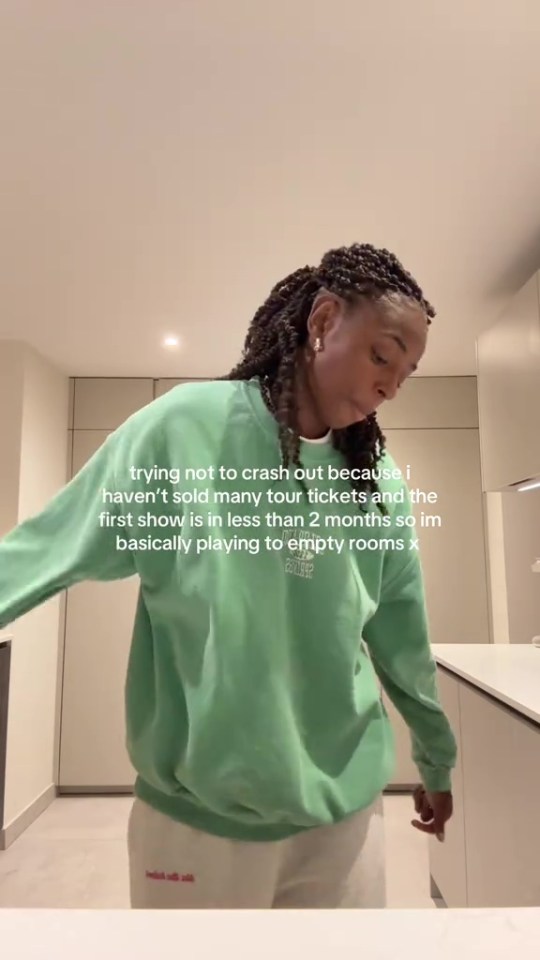 A woman with braided hair wearing a green sweatshirt and white pants, looking down while text on screen discusses not selling many tour tickets.