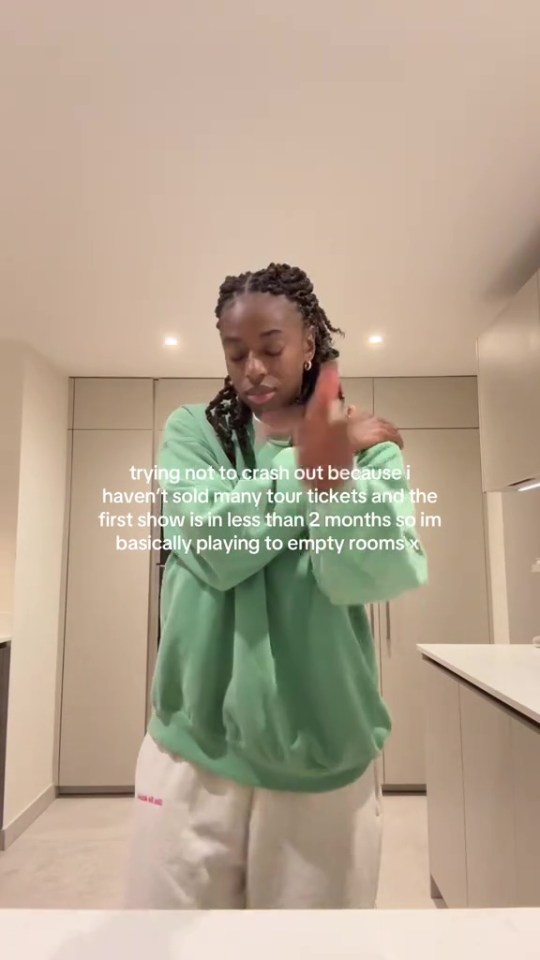 A woman in a green sweatshirt and white sweatpants gestures in a kitchen, with text overlay about her struggles to sell tour tickets.
