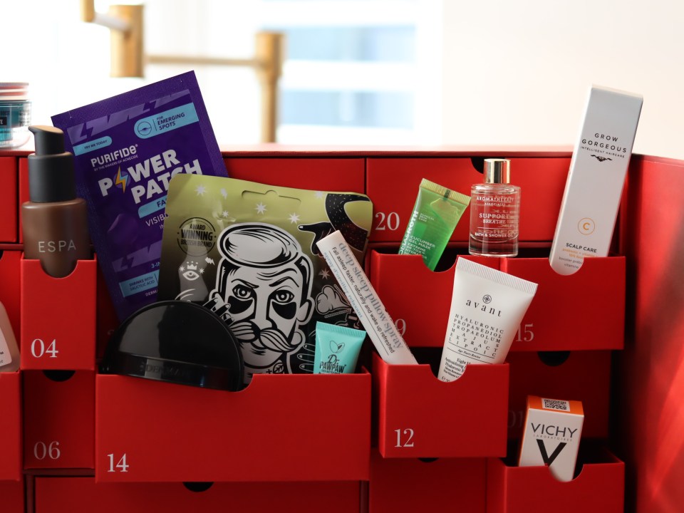 A red advent calendar filled with various beauty products in its opened drawers, including Purifide Power Patch, ESPA skincare, Dr. PawPaw lip balm, Aromatherapy Associates bath and shower oil, Grow Gorgeous Scalp Care, and Vichy skincare.