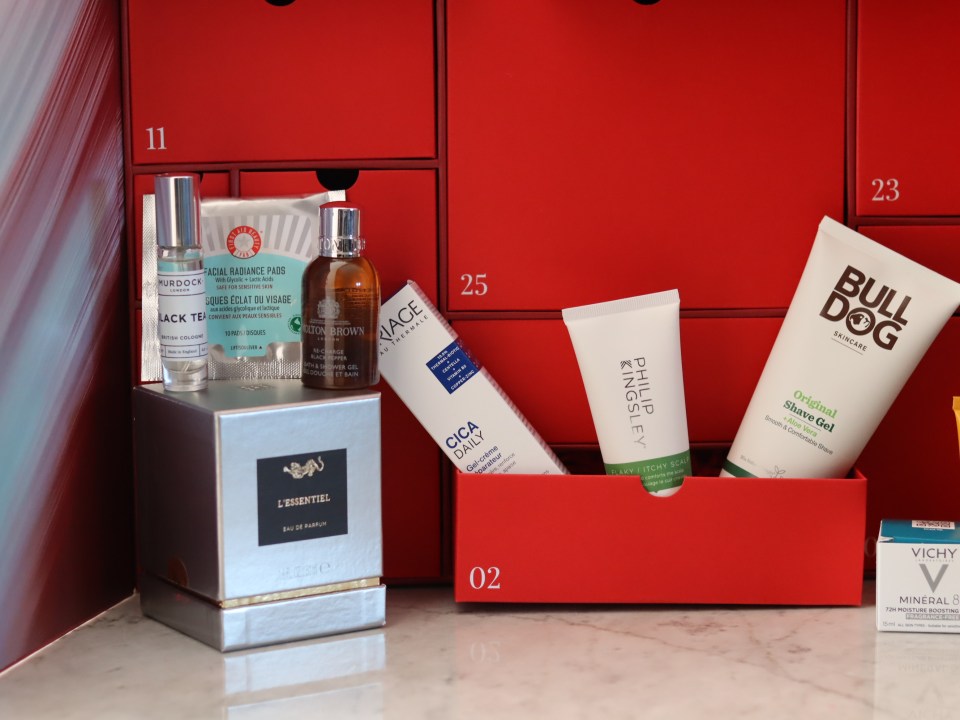 An assortment of beauty and skincare products including Murdock London cologne, Molton Brown bath and shower gel, First Aid Beauty facial radiance pads, Uriage Cica Daily gel-crème, Philip Kingsley flaky/itchy scalp treatment, Bull Dog Skincare shave gel, and Vichy Mineral 89 serum, arranged around red advent calendar drawers.