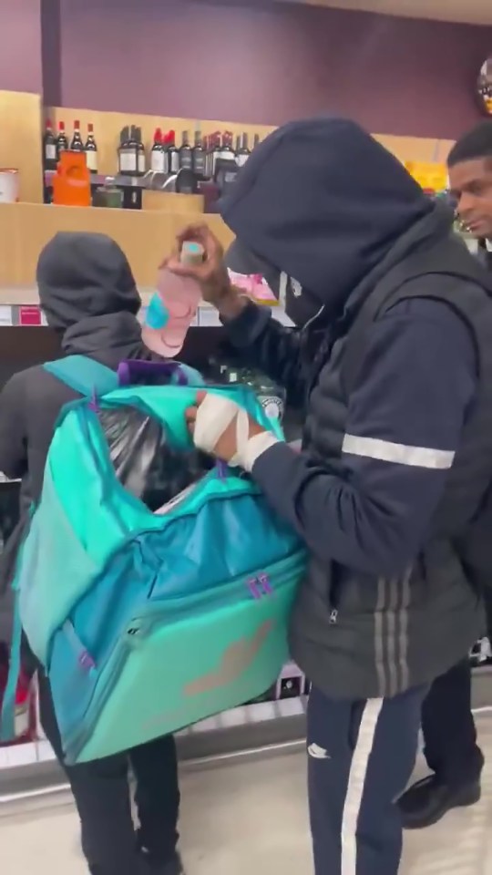 A video grab showing a man putting a bottle into a turquoise delivery bag while another man wearing a black hoodie stands behind him, in front of a shelf of bottled drinks.