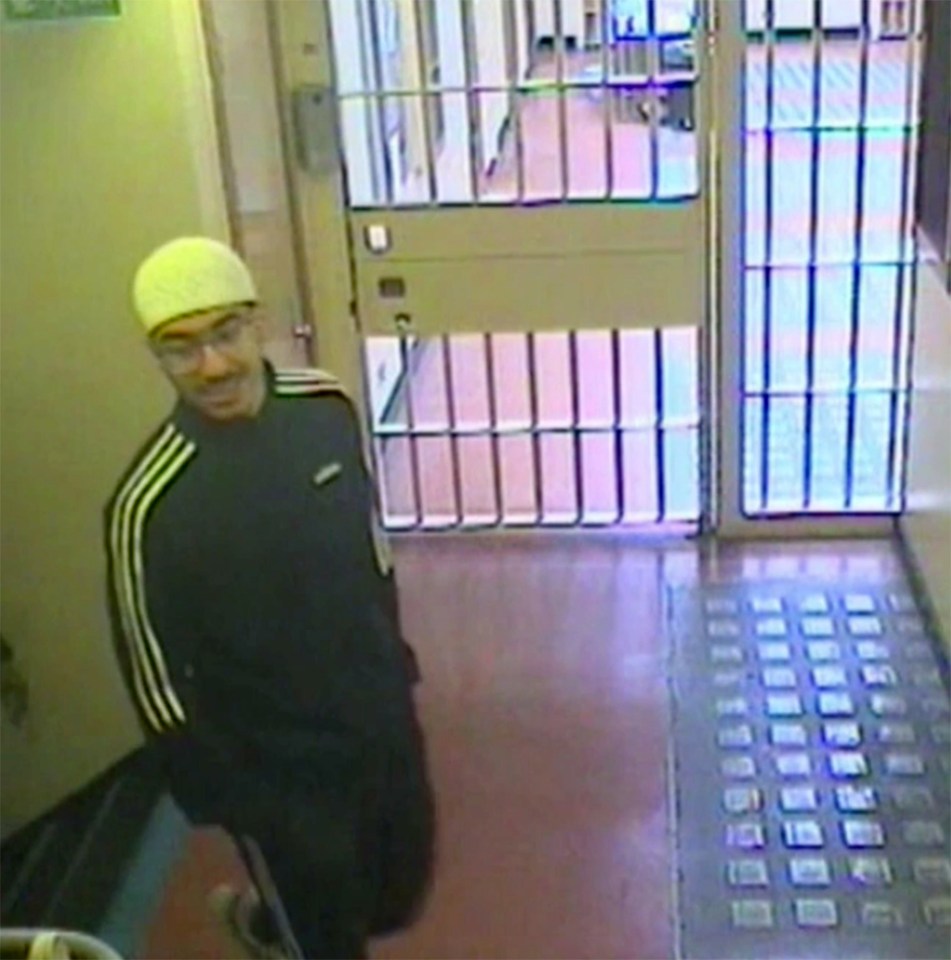 Hashem Abedi, wearing a yellow beanie, glasses, and a black Adidas jacket, smiles in a CCTV image from a prison hallway.