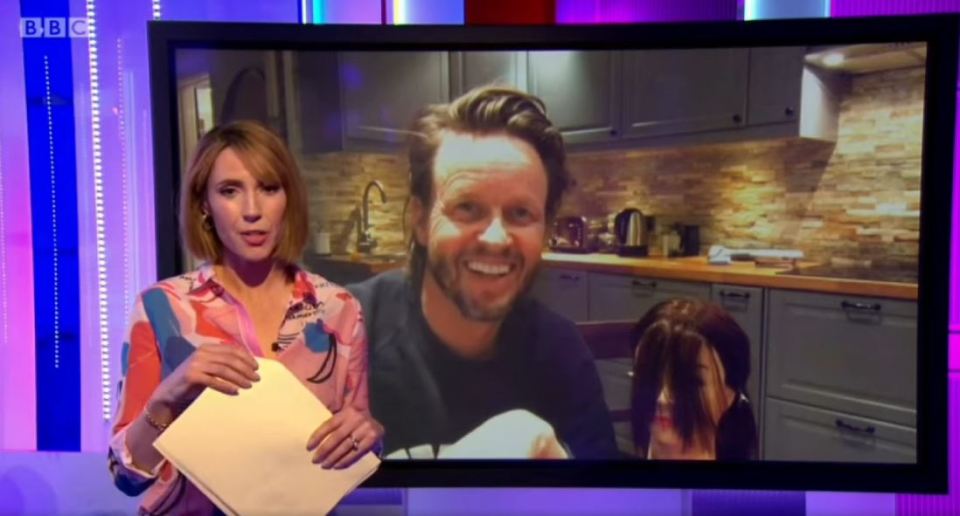 A woman on The One Show interviewing Michael Douglas on a video call demonstrating cutting and coloring hair at home.